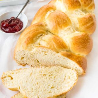 A loaf of braided Easter bread, with a slice cut off and lying in front of it on a white dish towel next to a small white bowl of strawberry jam with a spoon in it.