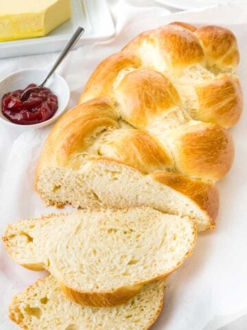 A loaf of braided Easter bread, with a slice cut off and lying in front of it on a white dish towel next to a small white bowl of strawberry jam with a spoon in it.