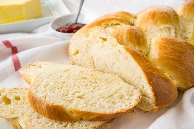 Close-up of a loaf of braided Easter bread, with two slices, cut off and lying in front of it on a white dish towel next to a small white bowl of strawberry jam with a spoon in it.