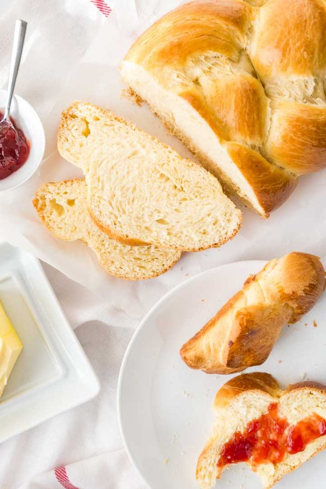 Top-down shot of braided Easter bread with two slices gut off and lying in front of it. Next to it, there\'s a white plate with slices of easter bread, a square plate with butter and a small white bowl of strawberry jam with a spoon in it.