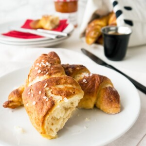 A white plate with pretzel croissants on a white tablecloth. There are more croissants, a stack of plates with red napkins and knives, a black cup of espresso and a black knife in the background.