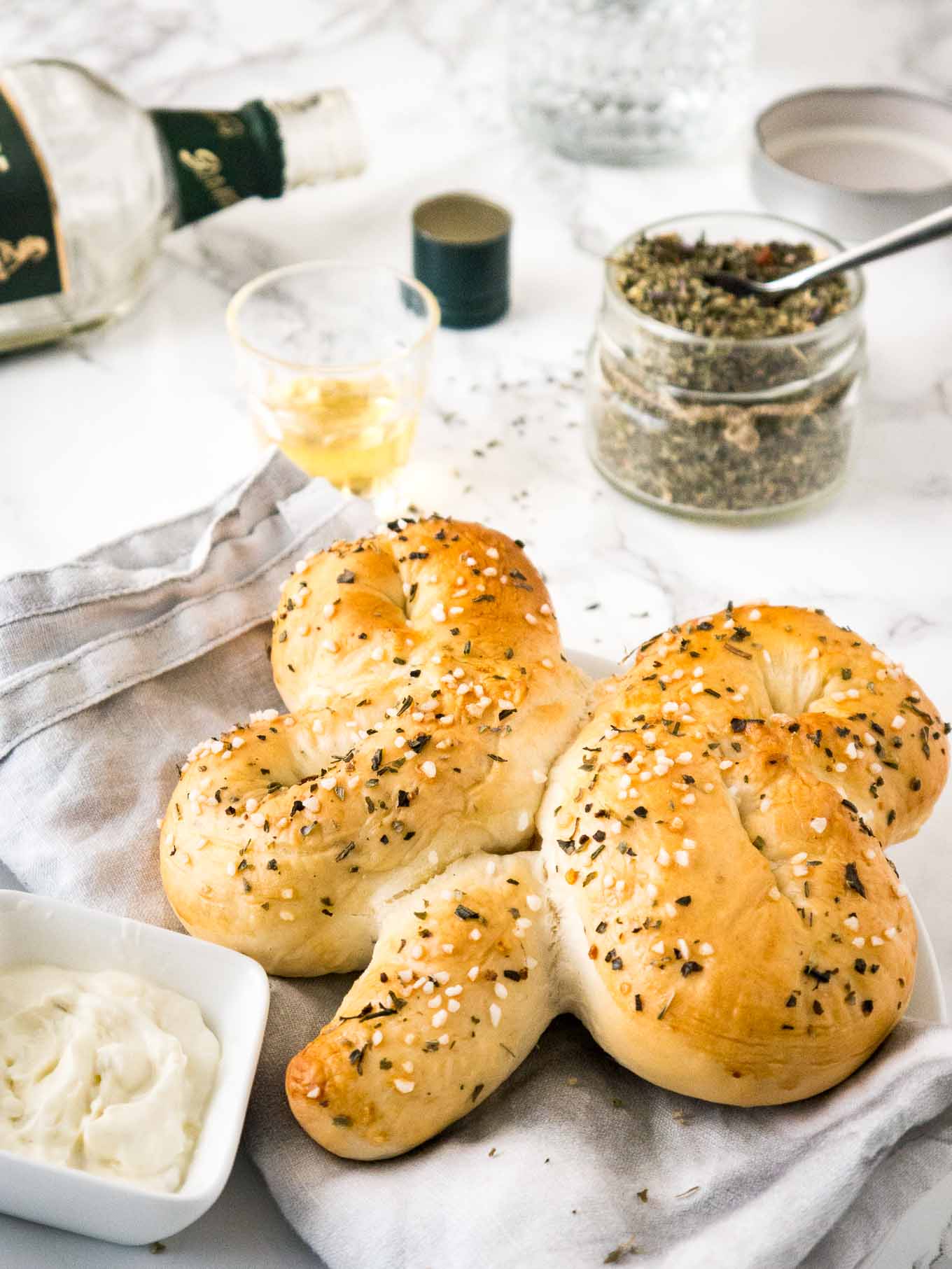 Close-up of a bread shaped like a shamrock on a grey dishtowel next to a small square bowl of dip on a plate. There\'s an empty bottle of whiskey, a shot of whiskey and a jar of spice mix in the background.