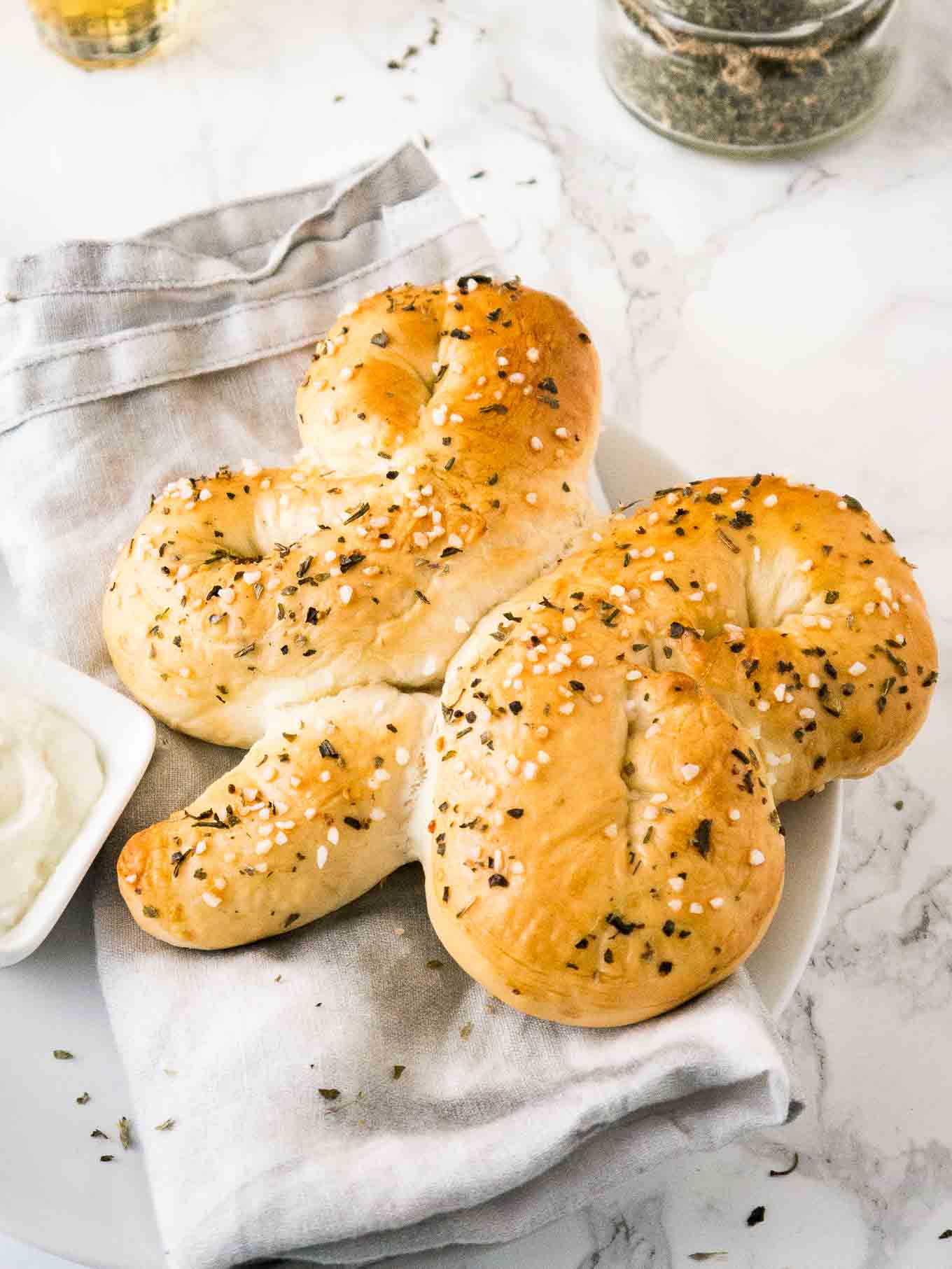 A bread shaped like a shamrock on a grey dishtowel next to a small square bowl of dip on a plate.