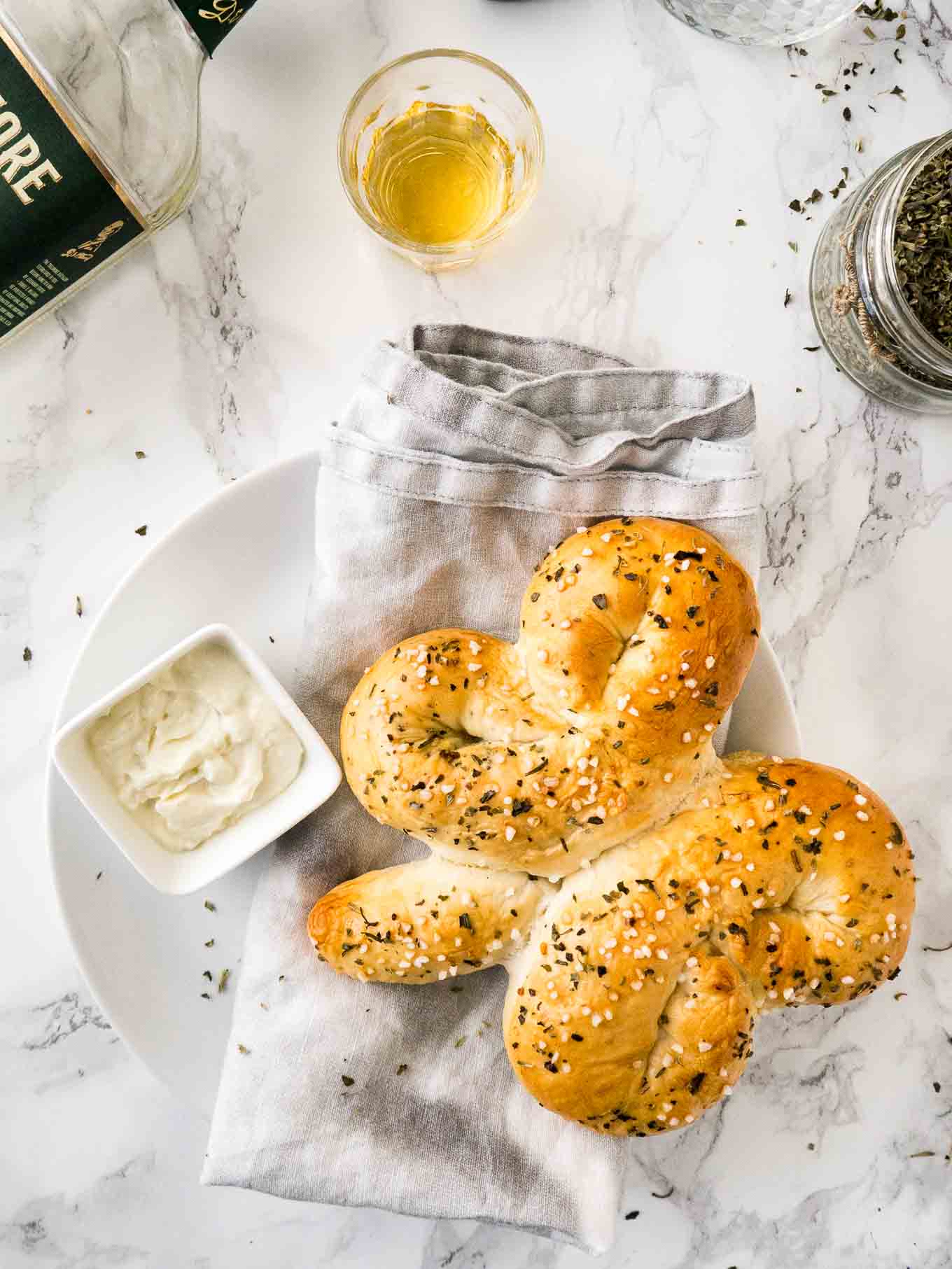 Top-down shot of a bread shaped like a shamrock on a grey dishtowel next to a small square bowl of dip on a plate.