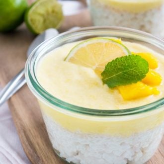 A jar with vegan breakfast mango rice pudding with coconut garnished with a slice of lime, mint and mango on a wooden cutting board. There's another jar of pudding in the background next to some spoons and halved limes.