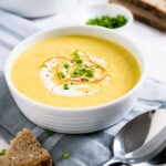 A white bowl of curried cream of corn soup, garnished with sour cream, chili threads and chives on a grey dishtowel. There's a piece of bread in front of it and a spoon next to it. Another bowl of soup, a small white bowl of chives and more bread is in the background.