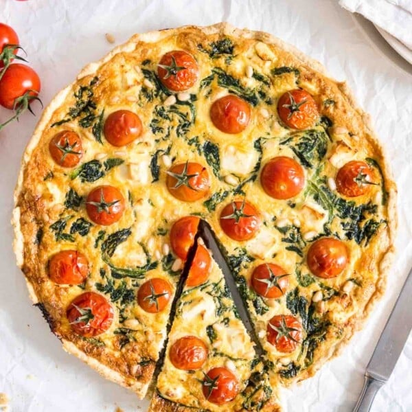 Spinach quiche with tomatoes sitting on a white dishtowel with a knife, a red and white dishtowel and some tomatoes next to it. A slice has been cut but is still in place.