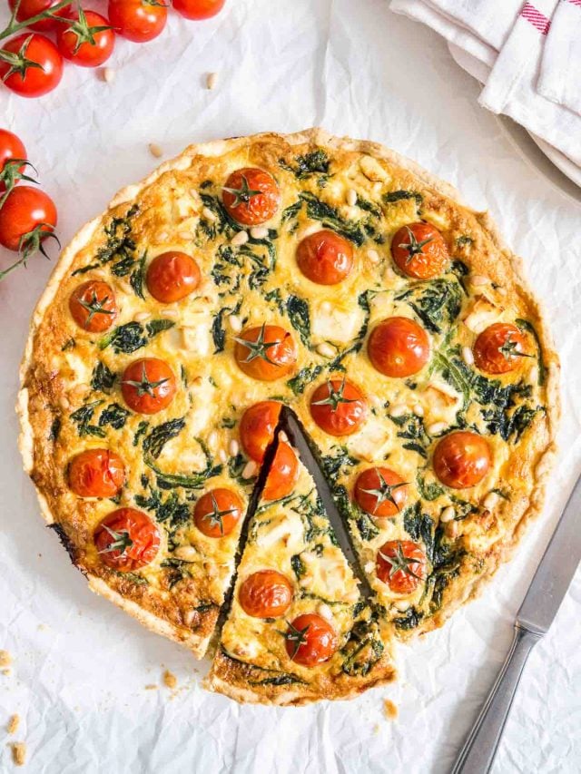 Spinach quiche with tomatoes sitting on a white dishtowel with a knife, a red and white dishtowel and some tomatoes next to it. A slice has been cut but is still in place.