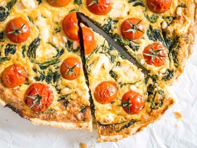 Close-up of spinach quiche with tomatoes sitting on parchment paper. A slice has been cut but is still in place.