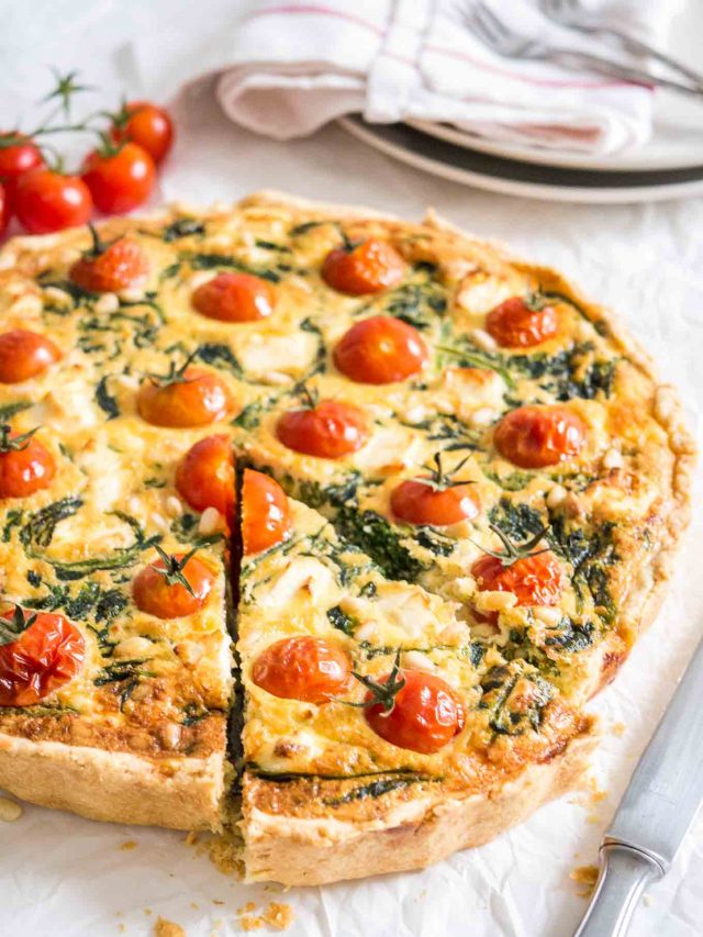 Close-up of spinach quiche with tomatoes sitting on a white dishtowel with a knife, a red and white dishtowel and some tomatoes next to it. A slice has been cut but is still in place.