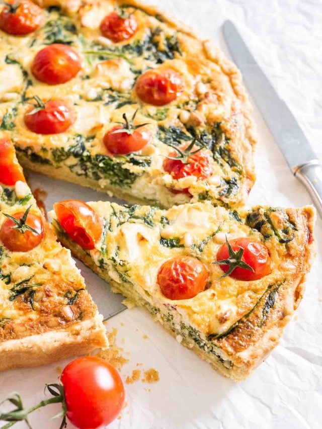 Close-up of spinach quiche with tomatoes sitting on a white dishtowel with a knife and some tomatoes next to it. A slice has been cut and pulled out slightly.