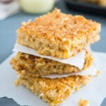 A stack of tropical coconut squares on a piece of parchment paper. There's a glass of sweetened condensed milk in the background next to a baking sheet with the rest of the squares.