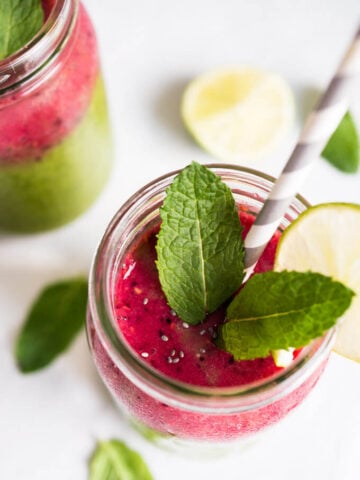 A jar of coconut water smoothie, with raspberries, baby spinach, chia, and mango, garnished with mint and a slice of lime with a straw in it. There's another glass of the smoothie an limes and mint next to it.