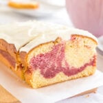A loaf of raspberry swirl pound cake sitting on a wooden cutting board lined with parchment paper. A slice has been cut off and is leaning against it. In the background, there's a pink cup and a stack of two plates with another slice on it.
