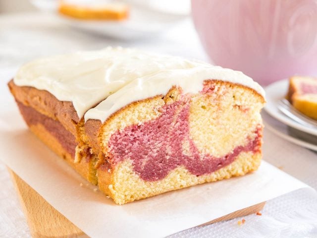 A loaf of raspberry swirl pound cake sitting on a wooden cutting board lined with parchment paper. A slice has been cut off and is leaning against it. In the background, there\'s a pink cup and a stack of two plates with another slice on it.