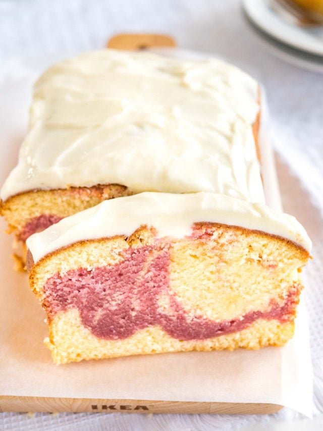 A loaf of raspberry swirl pound cake sitting on a wooden cutting board lined with parchment paper. A slice has been cut off and is leaning against it. In the background, there are two plates with another slice on it.