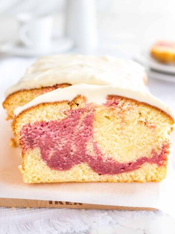 A loaf of raspberry swirl pound cake sitting on a wooden cutting board lined with parchment paper. A slice has been cut off and is leaning against it. In the background, there are two plates with another slice on it.