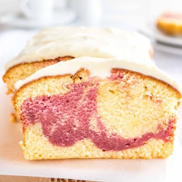A loaf of raspberry swirl pound cake sitting on a wooden cutting board lined with parchment paper. A slice has been cut off and is leaning against it. In the background, there are two plates with another slice on it.