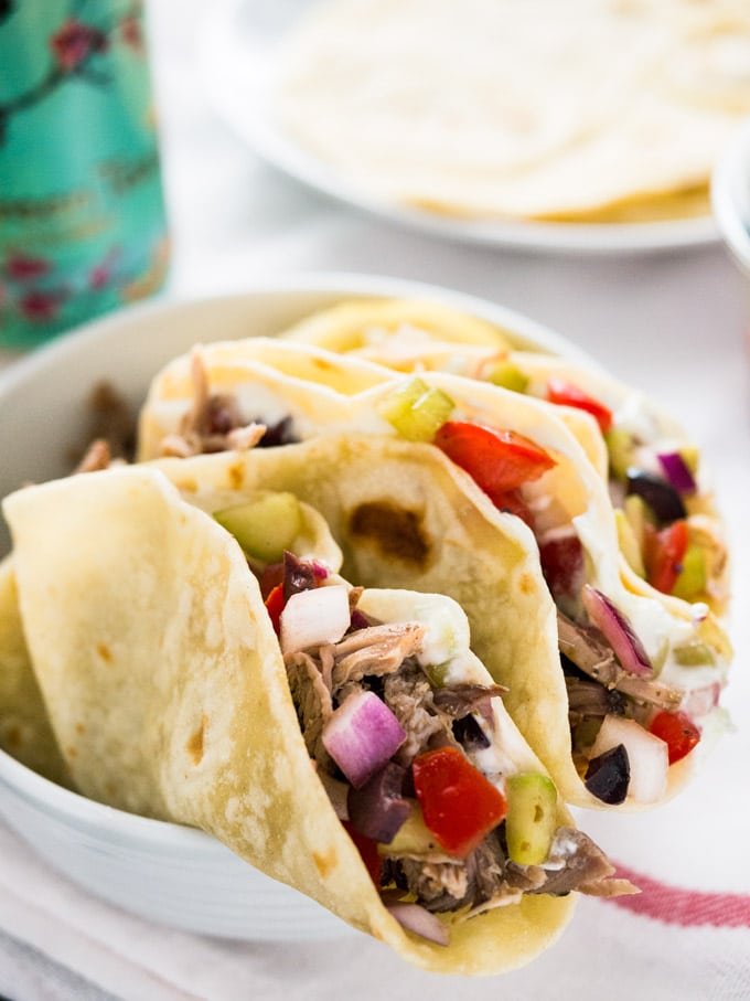 A white bowl greek style tacos, filled with greek style pulled pork, salad and tzatziki. There\'s a white plate of tortillas and a can of Arizona ice tea in the background.