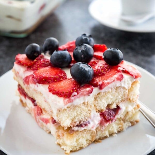 A white plate with a piece of strawberry tiramisu topped with blueberries. The rest of the tiramisu is in a glass baking dish in the background next to a white coffee cup.