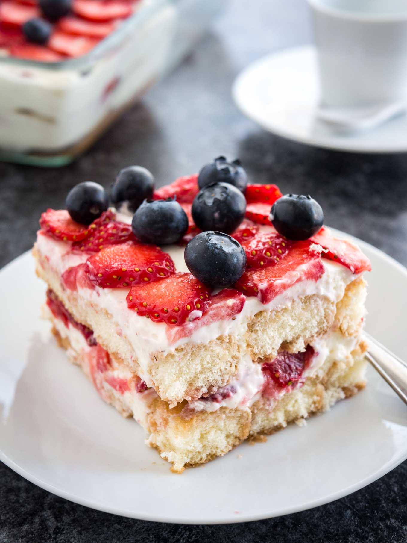 A white plate with a piece of strawberry tiramisu topped with blueberries. The rest of the tiramisu is in a glass baking dish in the background next to a white coffee cup.