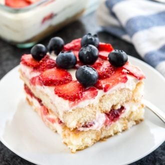 A white plate with a piece of strawberry tiramisu topped with blueberries. The rest of the tiramisu is in a glass baking dish in the background next to a white and blue dishtowel.