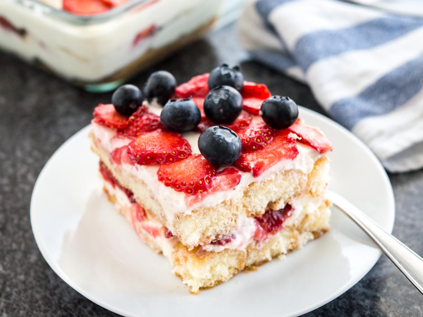 A white plate with a piece of strawberry tiramisu topped with blueberries. The rest of the tiramisu is in a glass baking dish in the background next to a white and blue dishtowel.