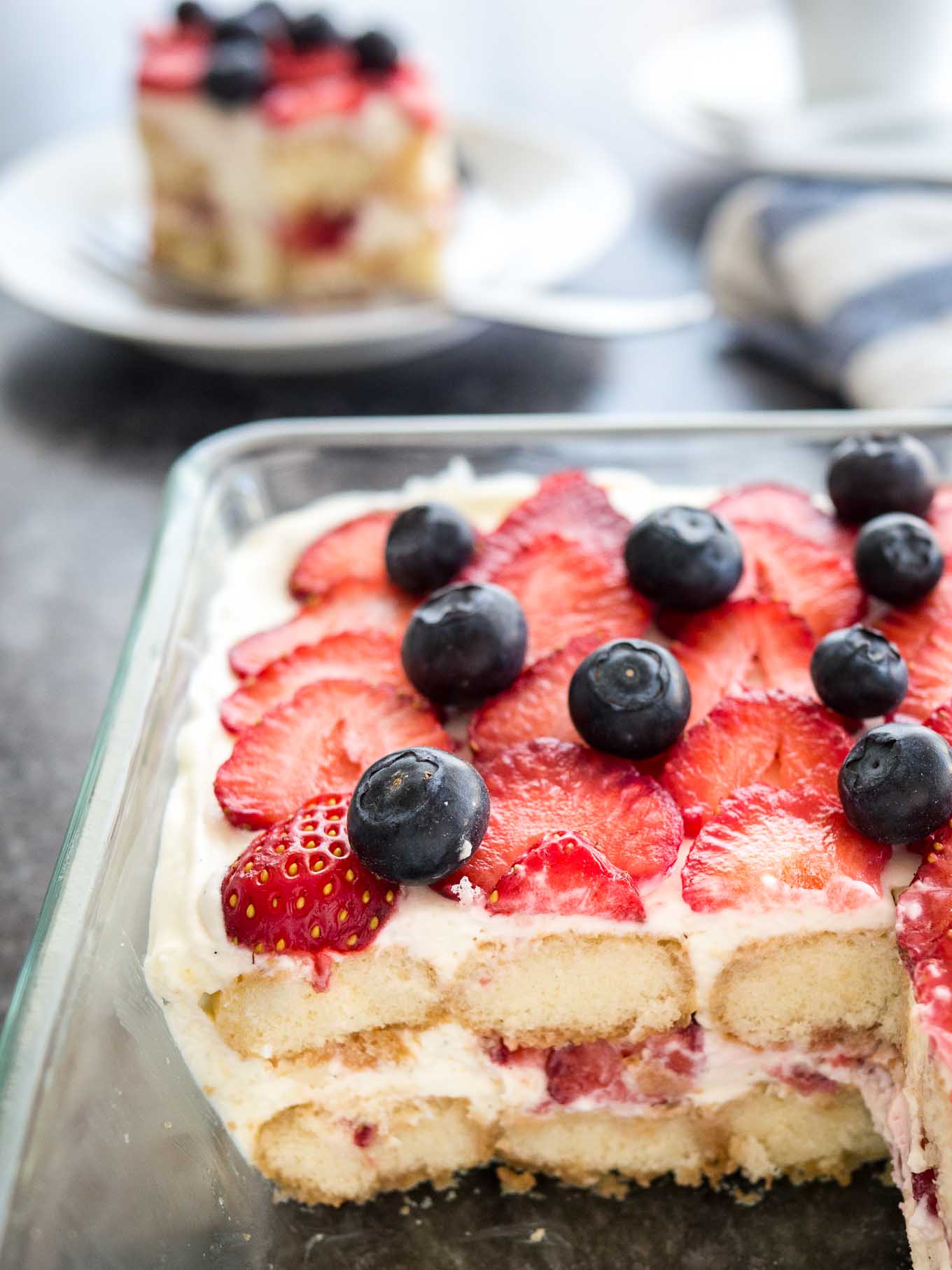 Strawberry tiramisu in a glass baking dish topped with blueberries. A piece of the tiramisu has been cut out and it is sitting on a white plate in the background next to a white coffee cup and a white and blue dishtowel.