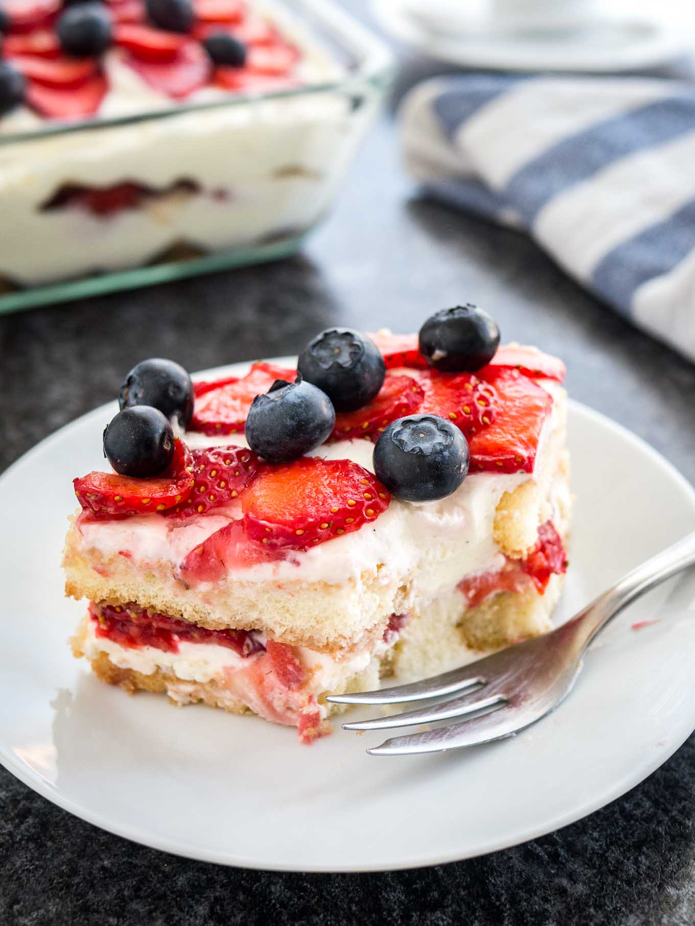 A white plate with a piece of strawberry tiramisu topped with blueberries. The rest of the tiramisu is in a glass baking dish in the background next to a white and blue dishtowel. A piece of the tiramisu has been removed and a fork is lying in front of it.