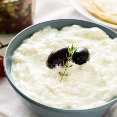 A slate grey bowl of Tzatziki, with to black olives and a sprig of thyme on a white dishtowel. There's a white plate of tortillas and a weck jar of greek salad in the background.