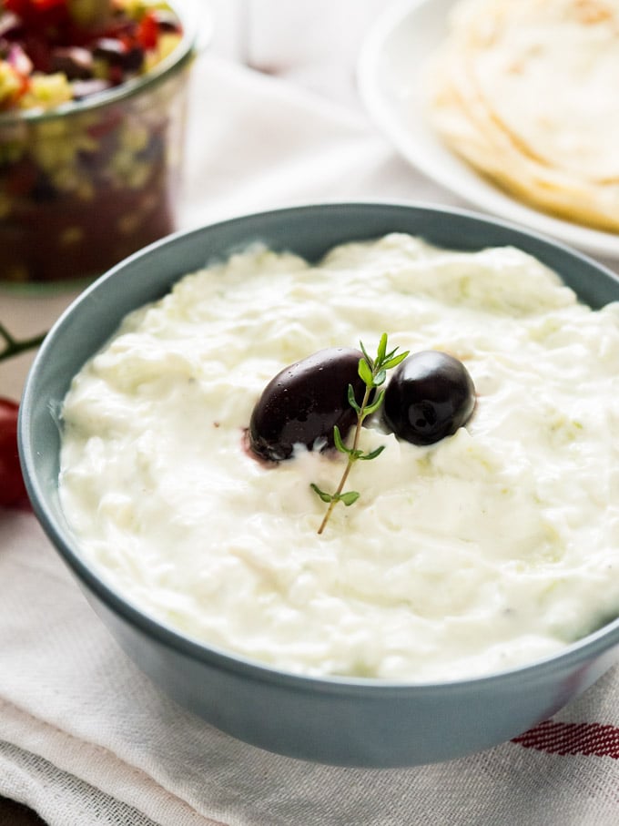 A slate grey bowl of Tzatziki, with to black olives and a sprig of thyme on a white dishtowel. There\'s a white plate of tortillas and a weck jar of greek salad in the background.