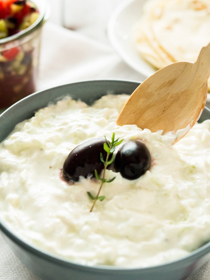 Close-up of a slate grey bowl of Tzatziki, with to black olives and a sprig of thyme with a wooden spoon in it on a white dishtowel. There\'s a white plate of tortillas and a weck jar of greek salad in the background.