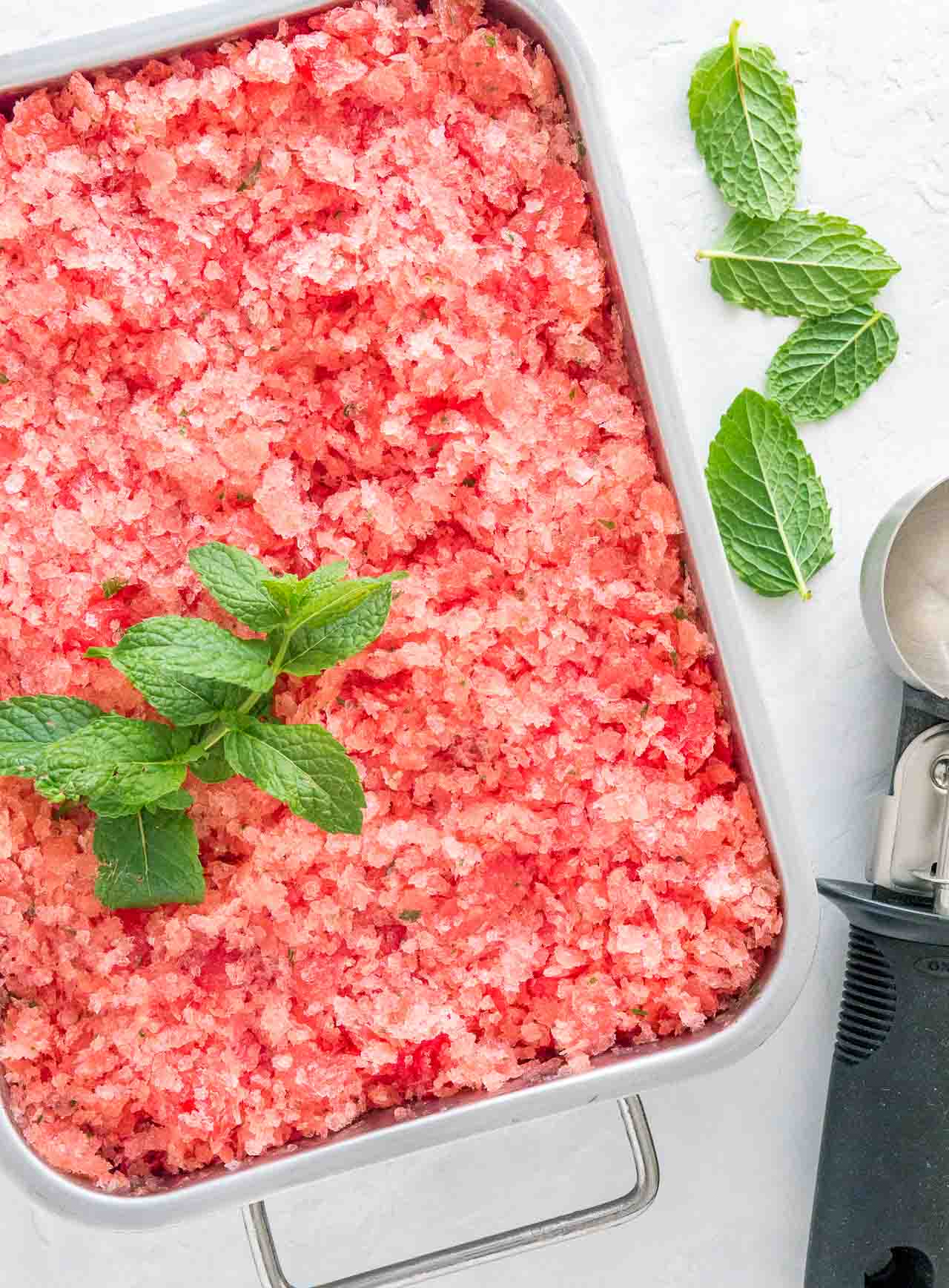 A stainless steel baking pan of pink watermelon granita garnished with mint with an ice cream scoop next to it.