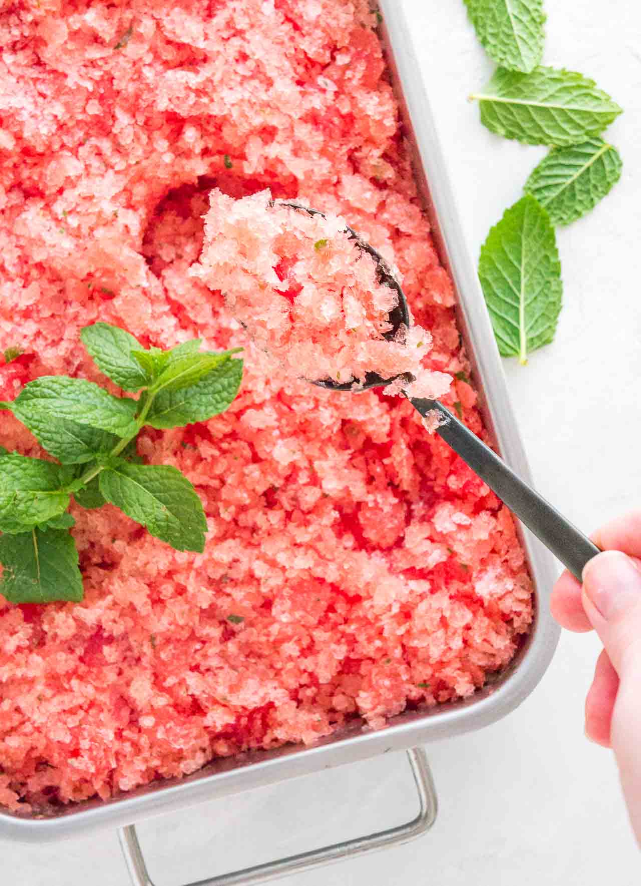 A stainless steel baking pan of pink watermelon granita garnished with mint. A hand is digging into the granita with a spoon.