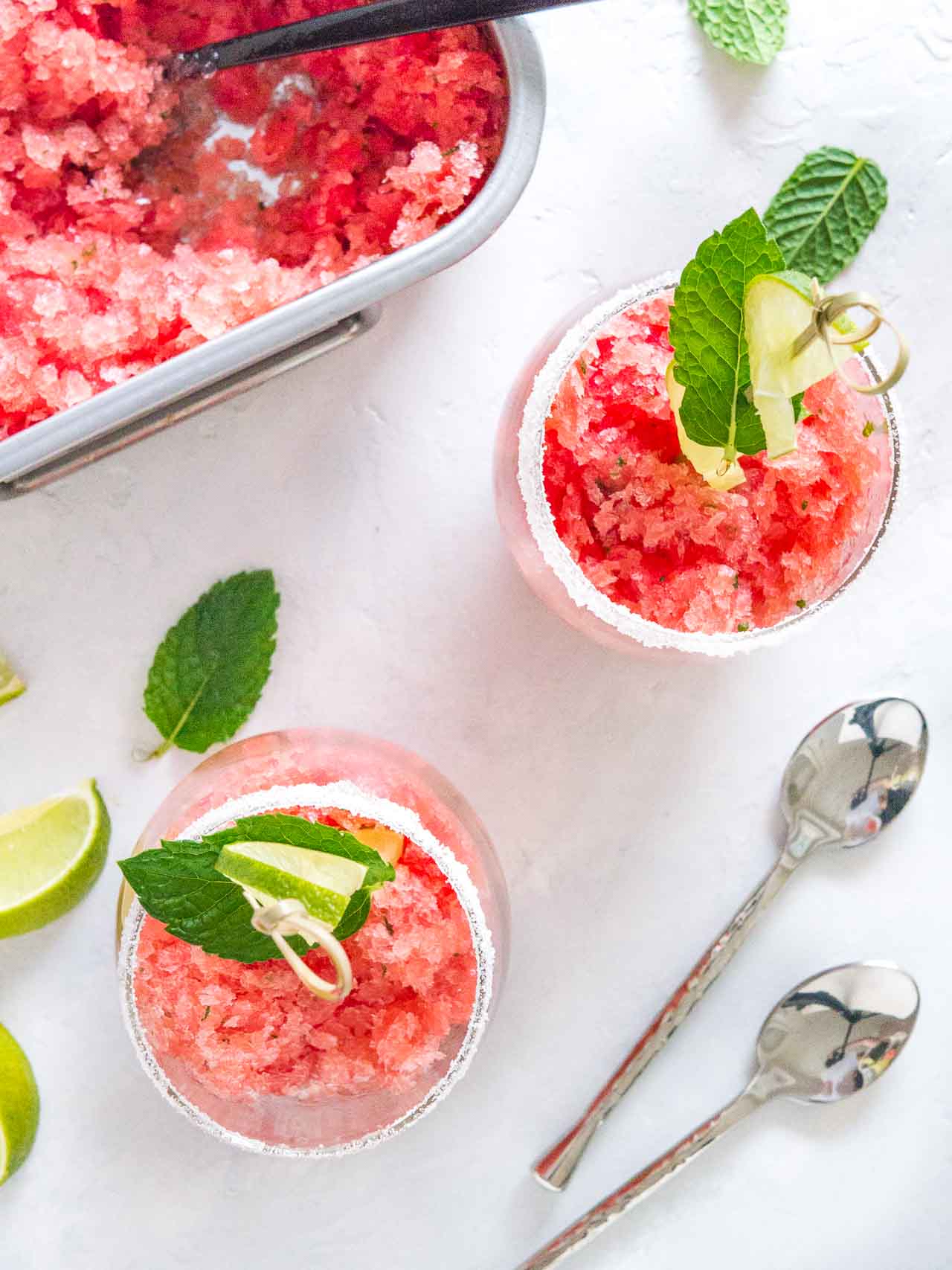 Top-down shot of two sugar-rimmed glasses of pink watermelon granita with skewers with lime and mint. There are spoons, lime wedges, mint and a stainless steel baking pan of granita next to it.