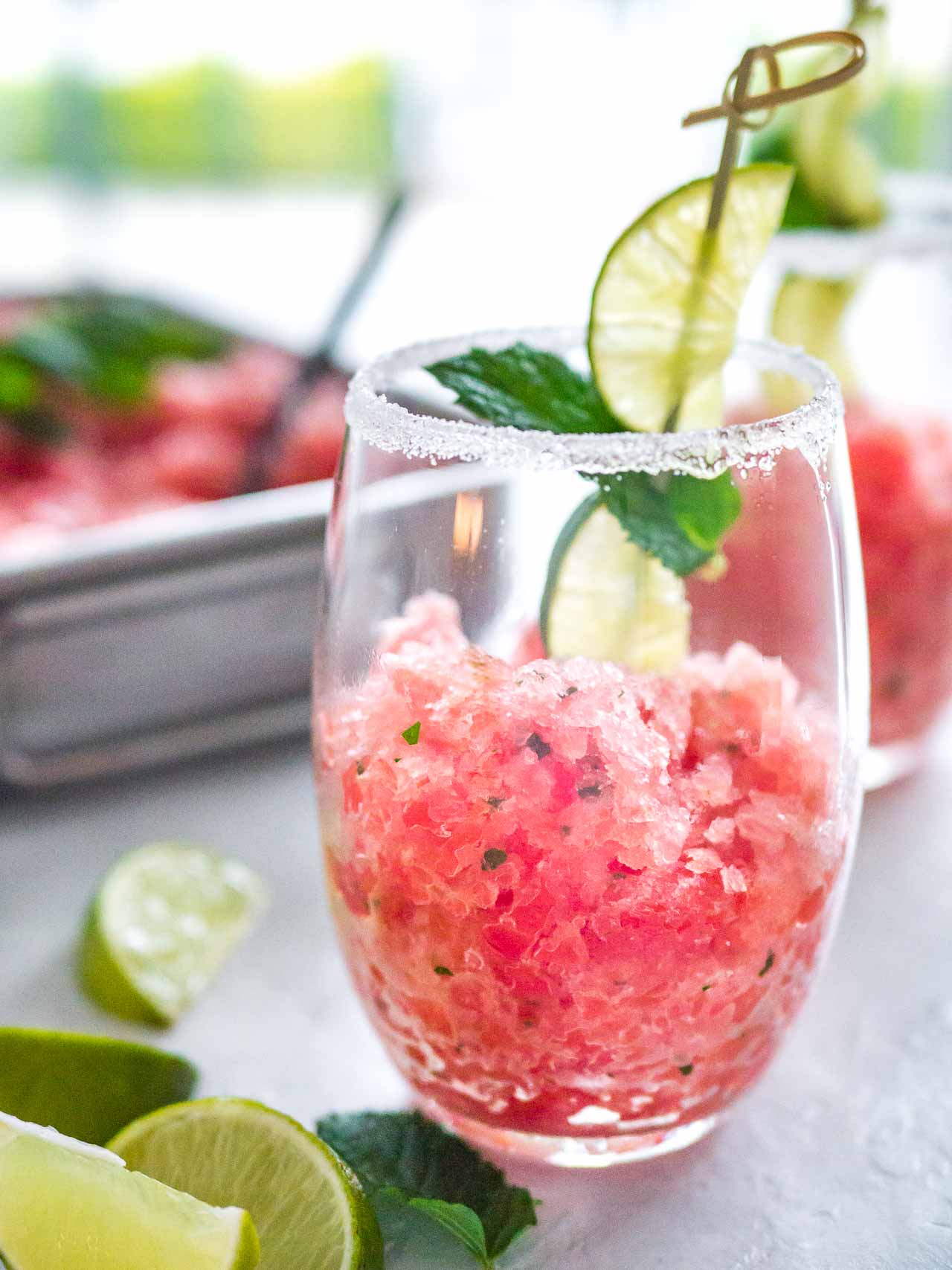 A sugar-rimmed glass with pink watermelon granita with a skewer of mint and lime on a grey surface. There\'s another glass of granita and a stainless steel baking pan with granita in the background and lime wedges next to it.