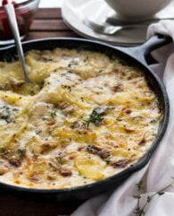 A cast-iron pan with gorgonzola potatoes au gratin wit a spoon digging into it on a wooden table. There's a white dishtowel next to it and a stack of plates with forks in the background.