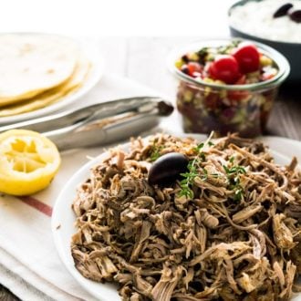 A plate of pulled pork, garnished with an olive and thyme sprigs on a white dishtowel with a red stripe. There's a jar of greek salad, a bowl of tzatziki, half a lime, some tongs and a plate of tortillas in the background.