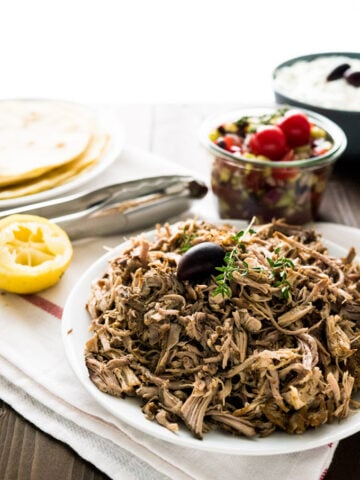 A plate of pulled pork, garnished with an olive and thyme sprigs on a white dishtowel with a red stripe. There's a jar of greek salad, a bowl of tzatziki, half a lime, some tongs and a plate of tortillas in the background.