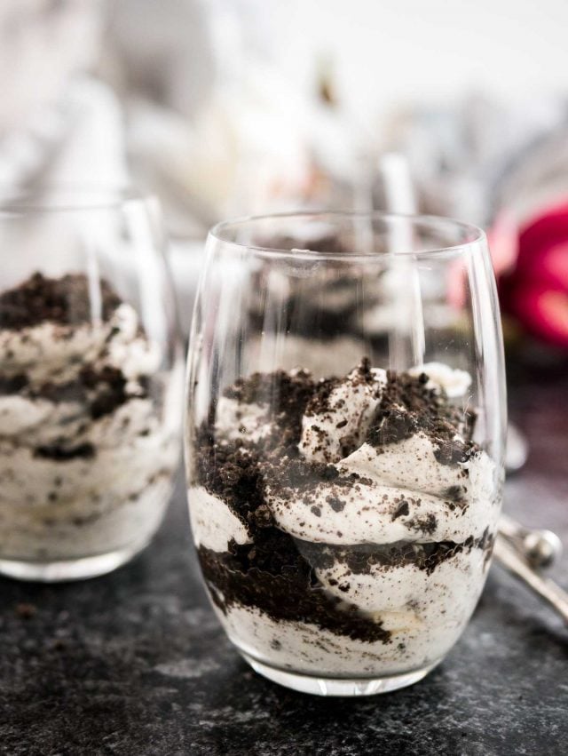 Two glasses of oreo cheesecake parfaits topped with oreo crumbs on a piece of parchment on a grey plate with a bronze spoon next to it.