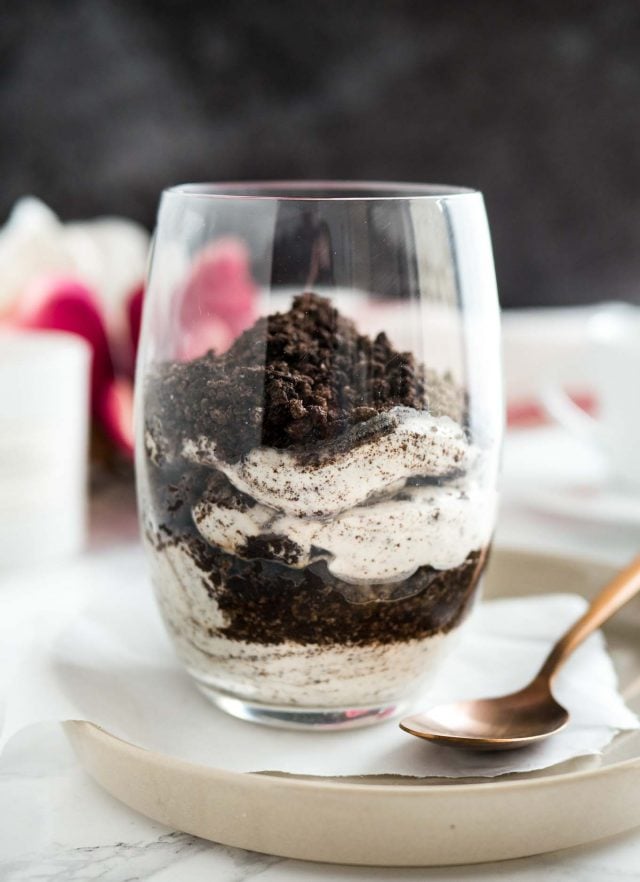 Close-up of a glass of oreo cheesecake parfaits topped with oreo crumbs on a piece of parchment on a grey plate with a bronze spoon next to it.