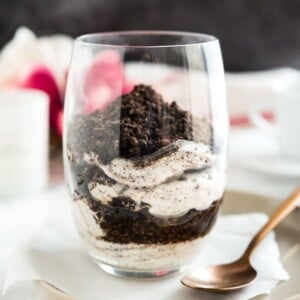 Close-up of a glass of oreo cheesecake parfaits topped with oreo crumbs on a piece of parchment on a grey plate with a bronze spoon next to it.