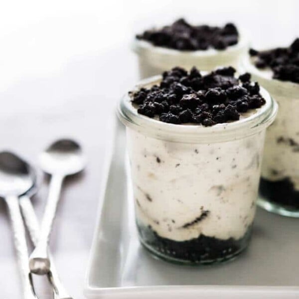 A rectangular white plate with 3 cylindrical weck jars with no-bake oreo cheesecake parfaits topped with oreo crumbs with spoons next to it.