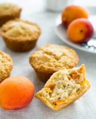 Skinny oatmeal apricot muffins on a grey tablecloth with some apricots. The frontmost muffin is halved and there's a white plate with apricots in the background.