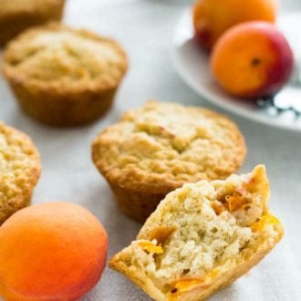 Skinny oatmeal apricot muffins on a grey tablecloth with some apricots. The frontmost muffin is halved and there's a white plate with apricots in the background.