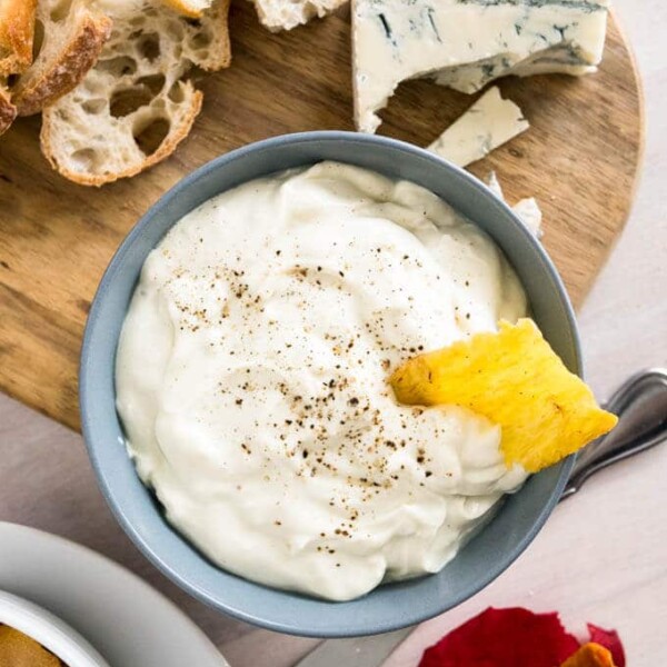 Top-down shot of a grey bowl of gorgonzola cream cheese spread on a wooden cutting board. There's a sliced loaf of bread and a piece of gorgonzola on the cutting board and a stack of white plates and rose petals next to it.