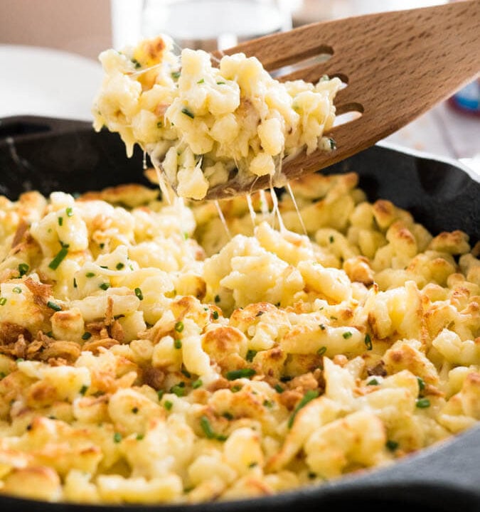 A cast-iron pan with cheese spaetzle. A slotted spatula is lifting out a portion.