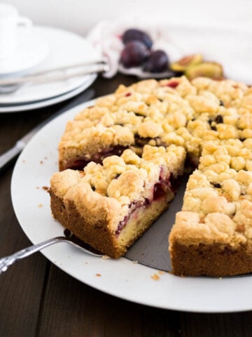 A plum cake with streusel topping on a springform bottom on a white plate on a wooden table. A cake server is lifting out a slice. There are stacked plates in the background.