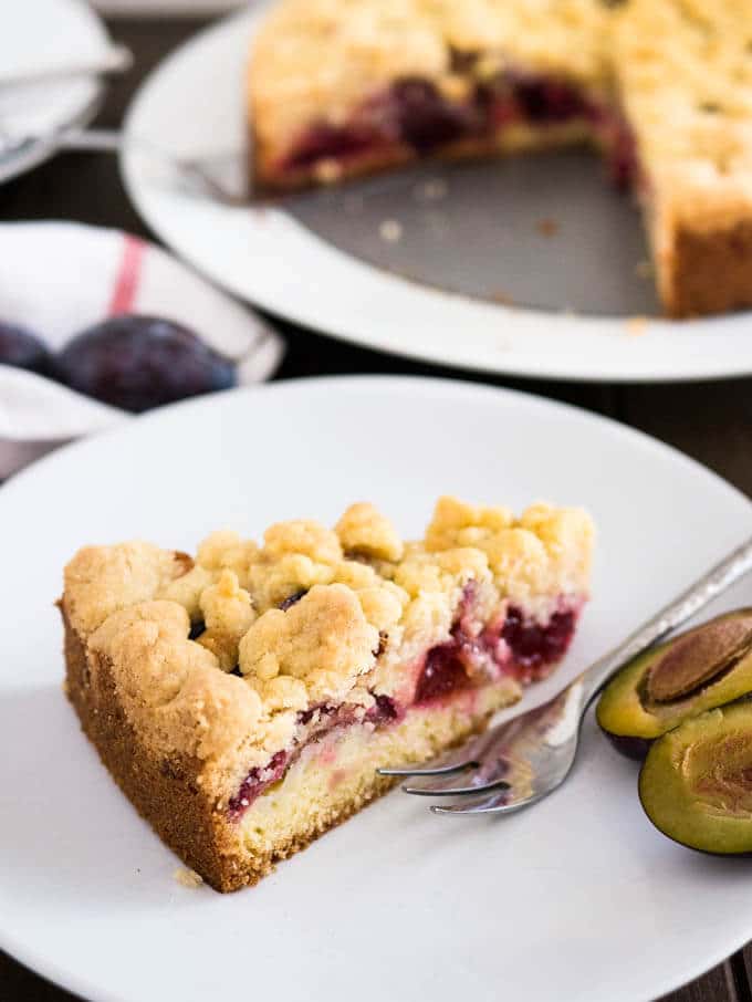 A slice of plum cake topped with streusel on a white plate with a fork and a halved plum. The whole cake is in the background.
