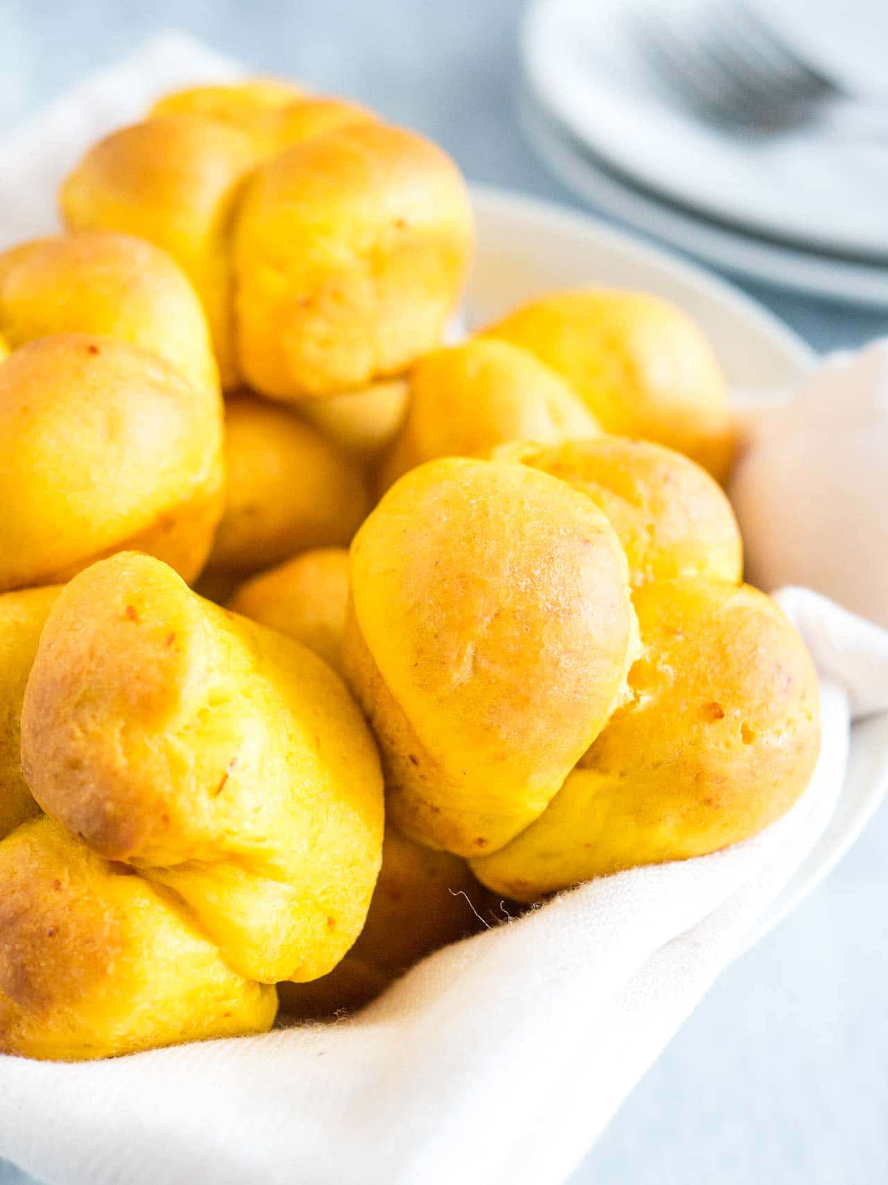 Close-up of pumpkin dinner rolls with a white dishtowel on a white plate with a stack of white plates in the background.